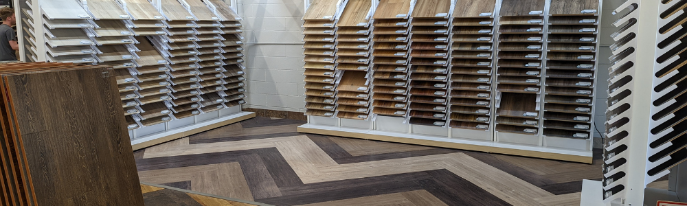 Bloomington Carpet One display of waterproof flooring samples arranged on wall racks, with multiple wood-look and tile-look panels laid out on the showroom floor under bright indoor lighting.